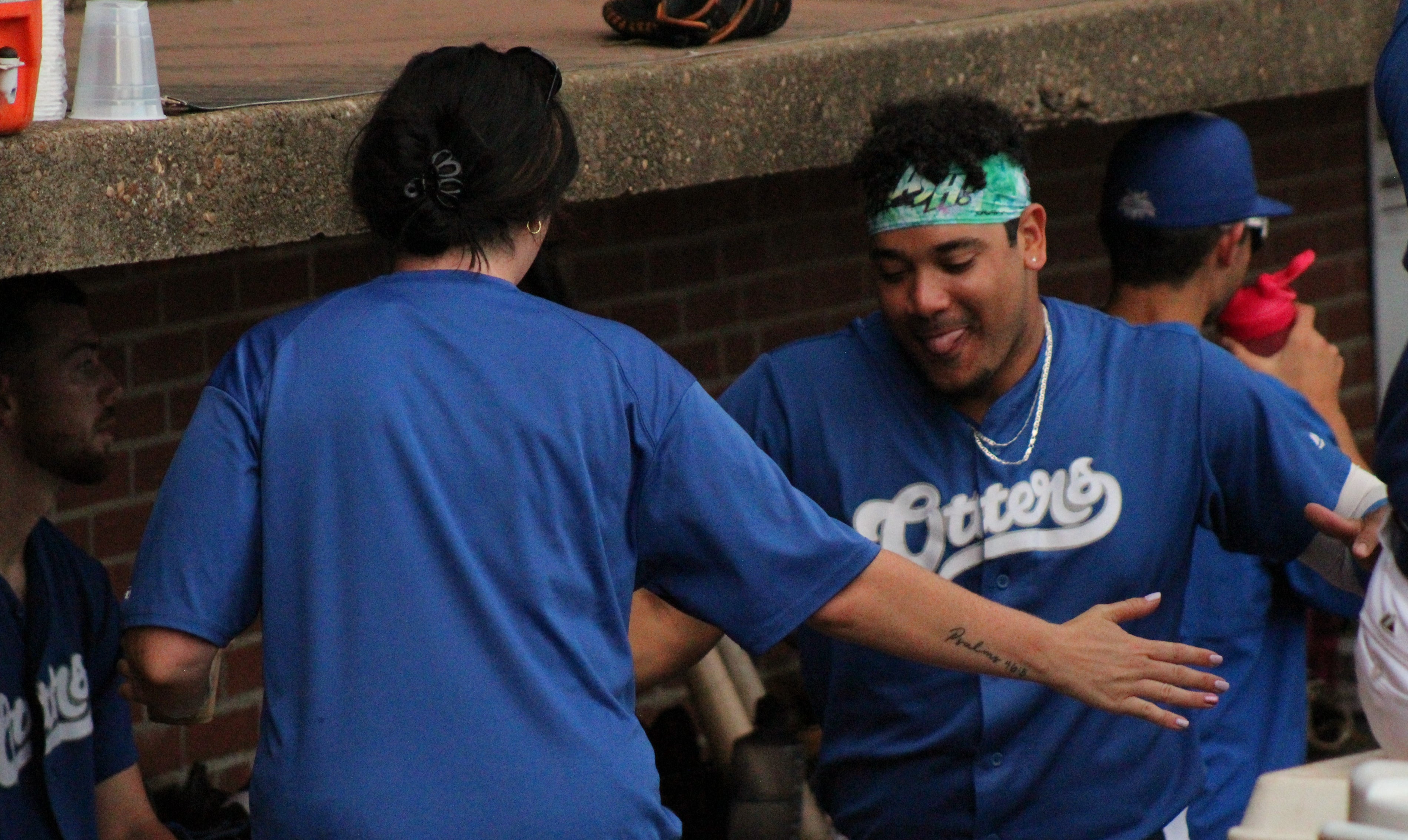 A White woman and a man with medium skin share a high-five in a baseball bench area. Both wear blue “Otters” shirts, and the man smiles while wearing a green headband. Other players sit in the background.