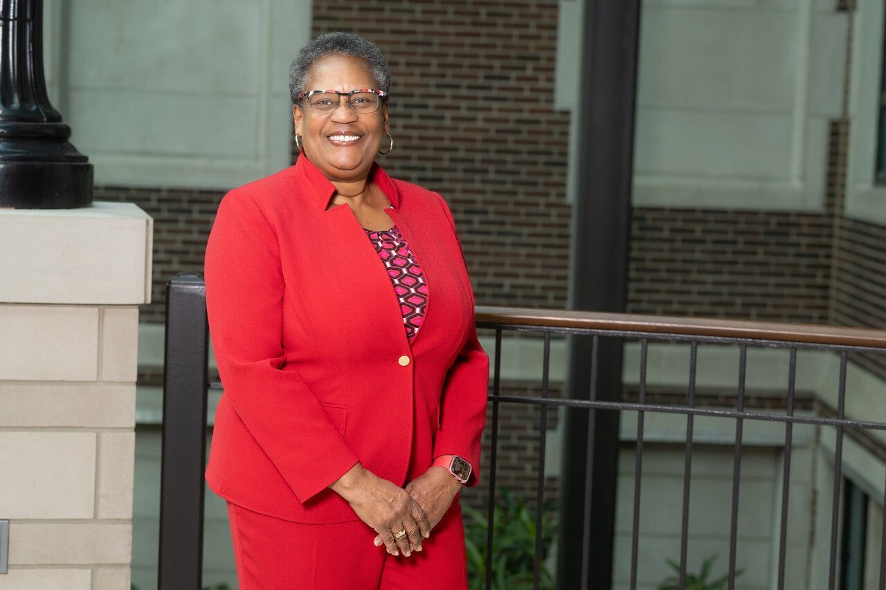 A smiling Black woman wearing glasses and a bright red suit stands indoors with her hands clasped. A brick wall and railing appear in the background.