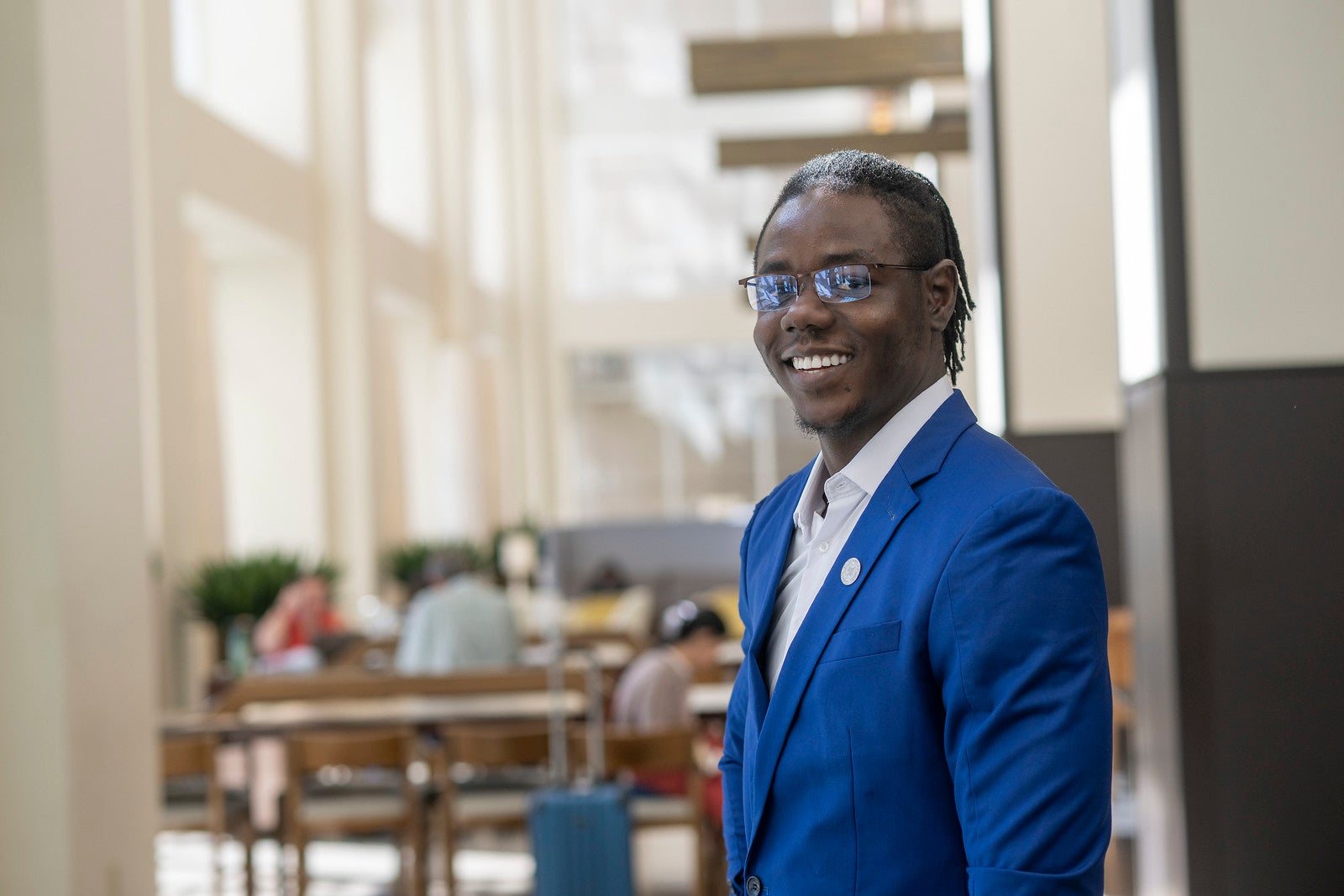 A Black man with braided hair, wearing a bright blue suit over a white shirt and glasses, stands in a modern, sunlit building with people seated in the background.