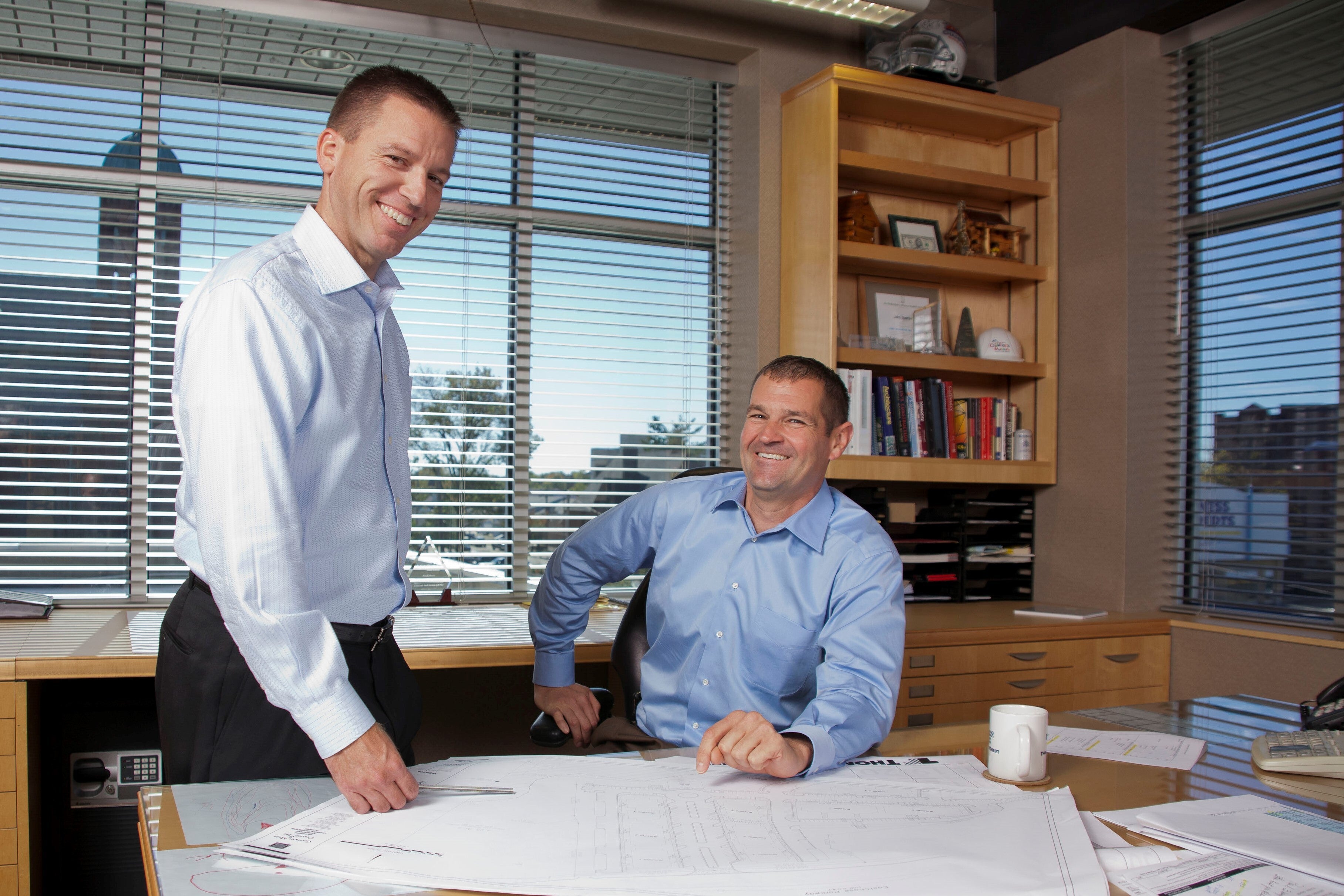 Two White men wearing light blue dress shirts work together in an office, with one standing and pointing at papers on the desk while the other sits smiling. Large windows and bookshelves are visible in the background.