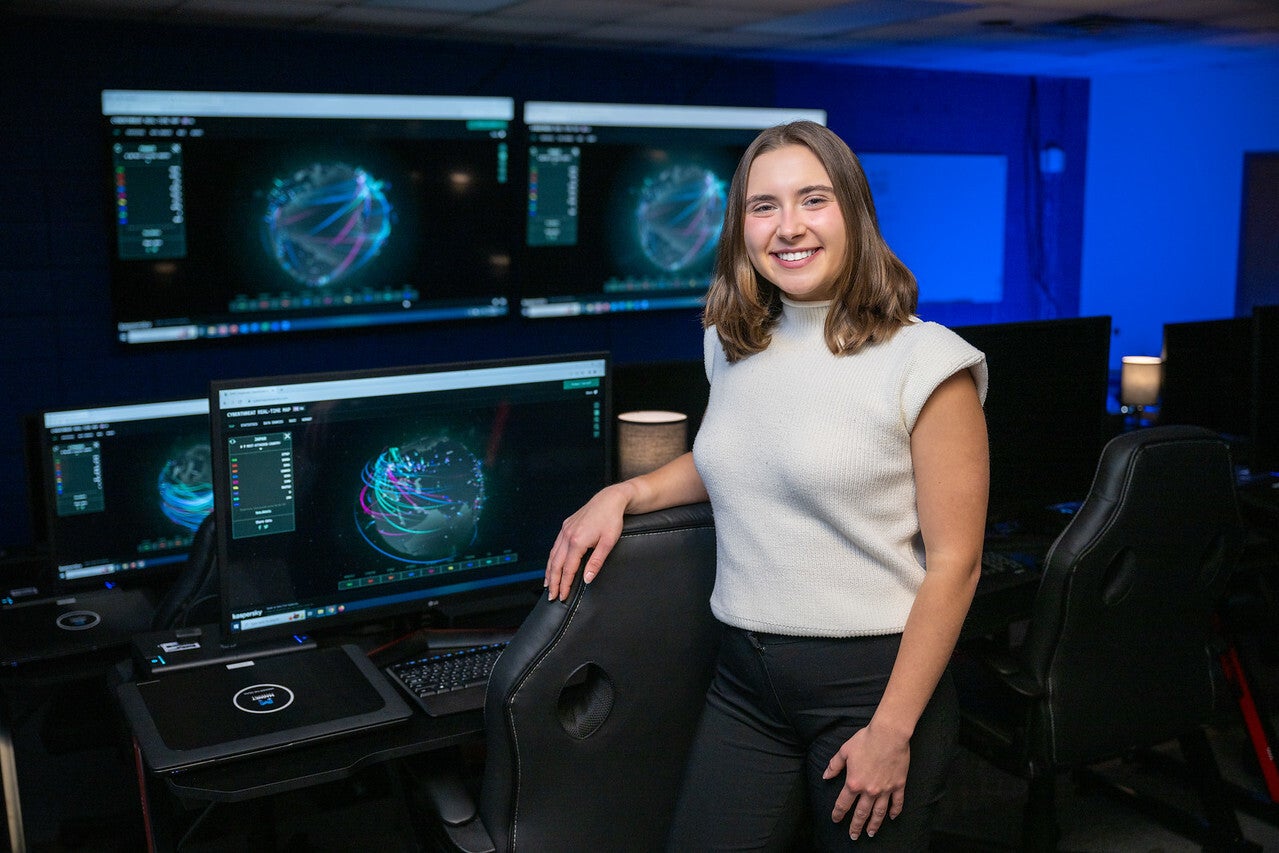 A young White woman with shoulder-length brown hair, wearing a white sleeveless top and black pants, stands smiling in a computer lab with multiple large monitors displaying colorful digital maps. She rests one hand on a chair in a dimly lit, blue-toned room.