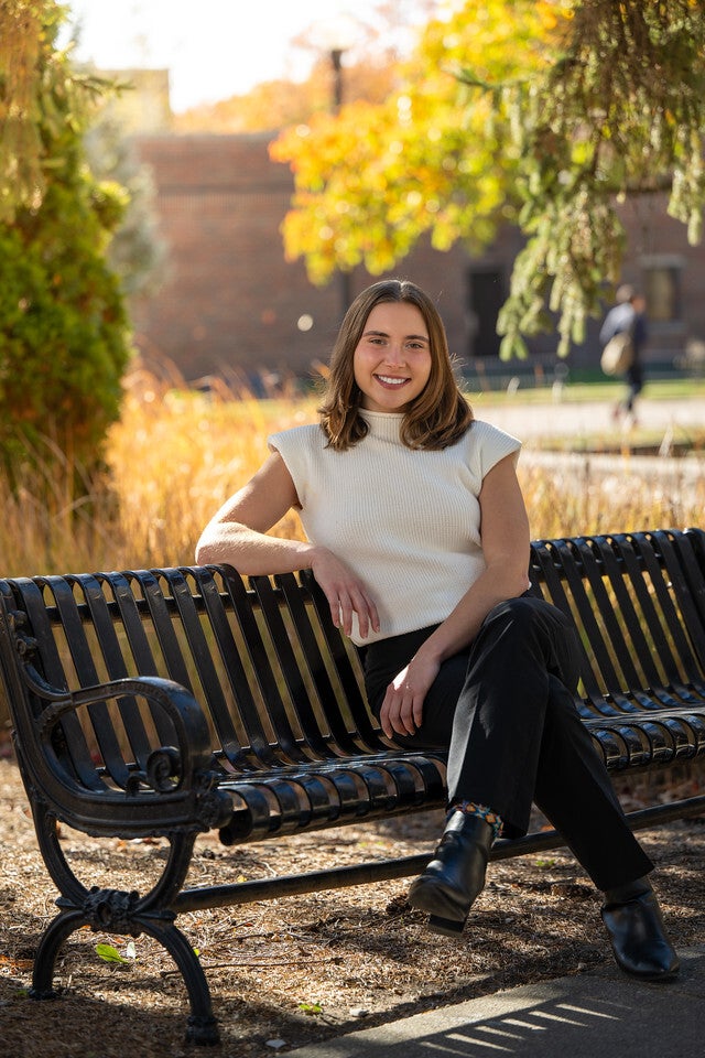 A young White woman with shoulder-length brown hair sits smiling on a black metal bench outdoors, wearing a white sleeveless top and black pants. Trees with yellow leaves and a brick building are visible in the background.