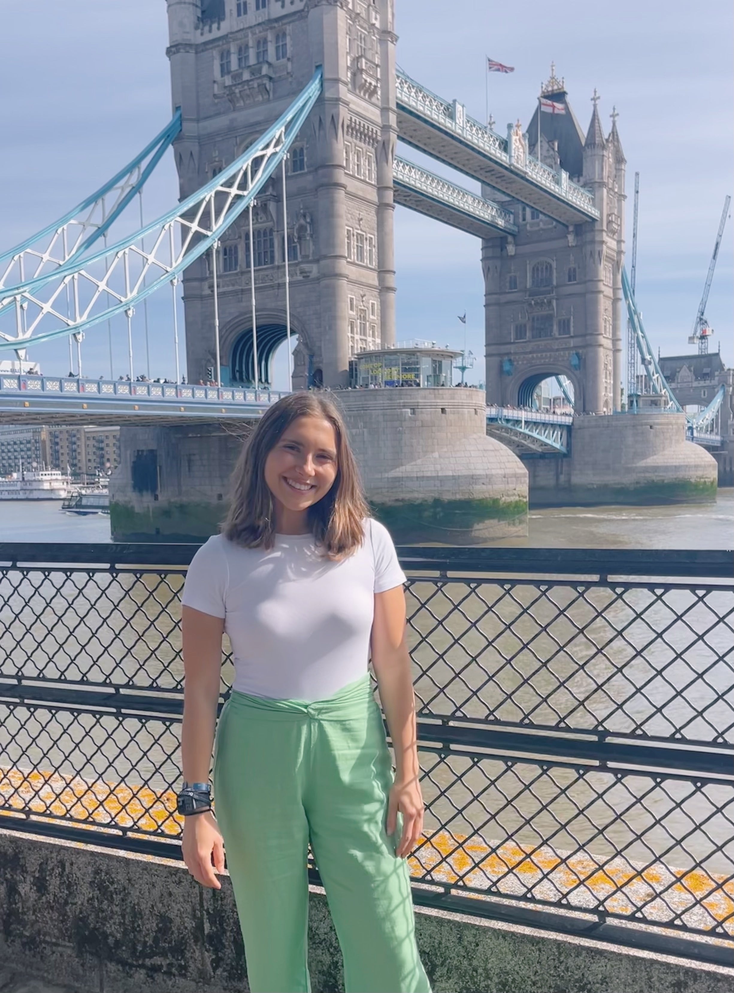 A young White woman with shoulder-length brown hair, wearing a white short-sleeve top and light green pants, stands smiling by a black metal railing near a river, with a large bridge visible in the background.