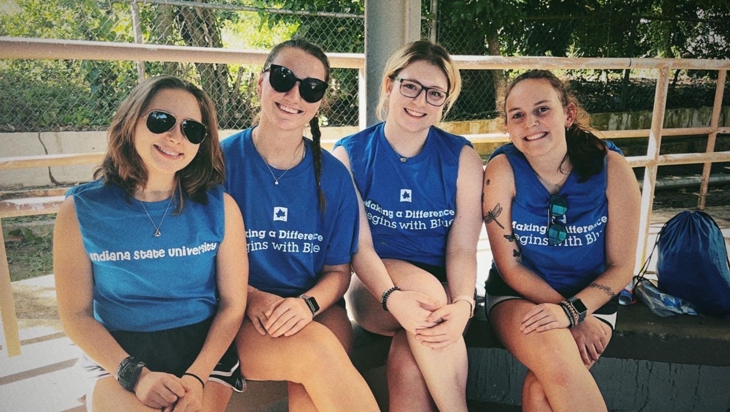 Four young White women sit side by side on a bench outdoors, wearing blue sleeveless shirts with “Indiana State University” and “Making a Difference Begins with Blue” text on the front. Two wear sunglasses. A fence, trees, and a blue drawstring bag are visible in the background.