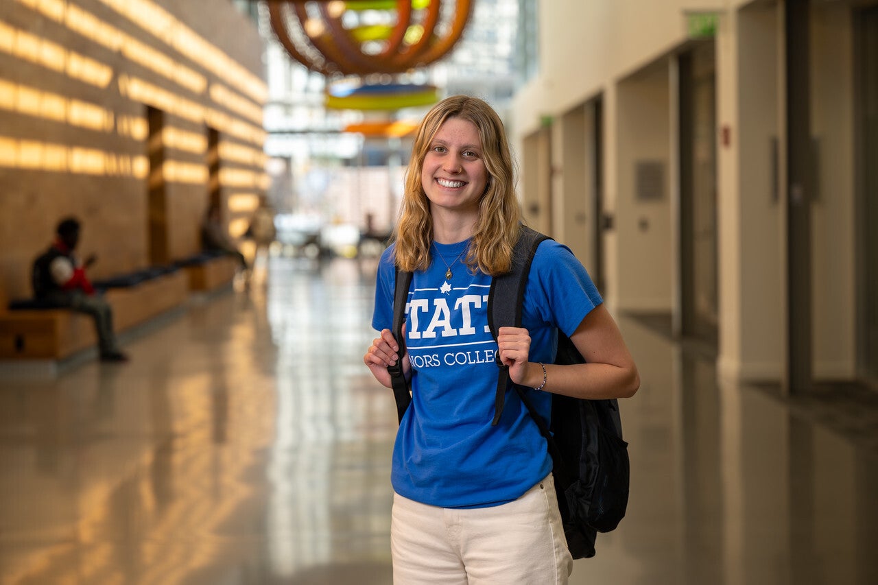 A young White woman with blonde hair stands smiling in a bright hallway, wearing a blue “STATE Honors College” T-shirt, light pants, and a black backpack. Warm sunlight and colorful ceiling art are visible in the background.