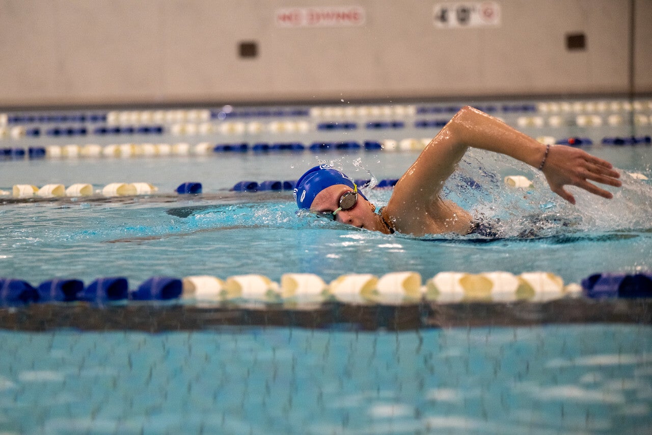 A female swimmer in a blue cap and goggles performs the freestyle stroke in an indoor pool, her head turned slightly as her arm slices through the water between blue and white lane dividers.