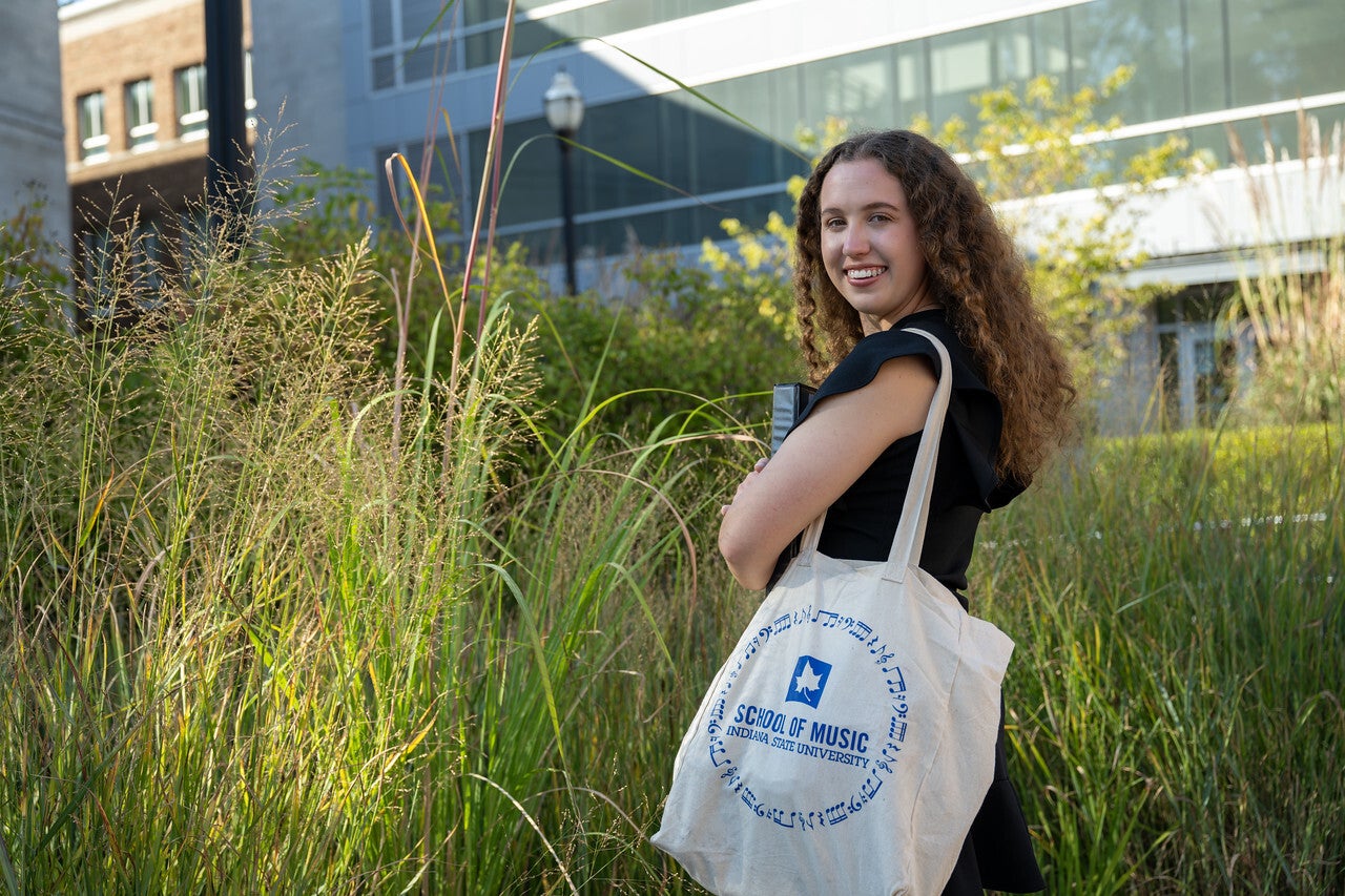 A white woman stands in tall grass near a modern building, smiling over her shoulder while holding a tote bag and folder. The tote bag reads “School of Music, Indiana University.”