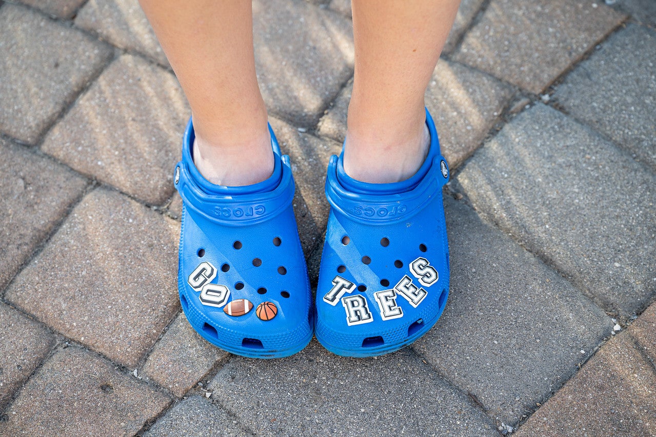 A close-up view of a person wearing bright blue Crocs decorated with white letter pieces that spell out “GO TREES,” along with small football and basketball shapes. The person stands on a tan brick walkway.