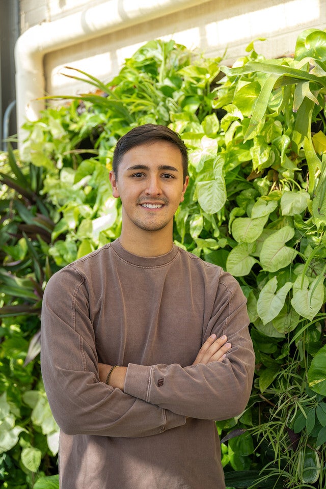 A young man with short dark brown hair, wearing a brown long-sleeve shirt, stands smiling with his arms crossed in front of a lush green plant wall.