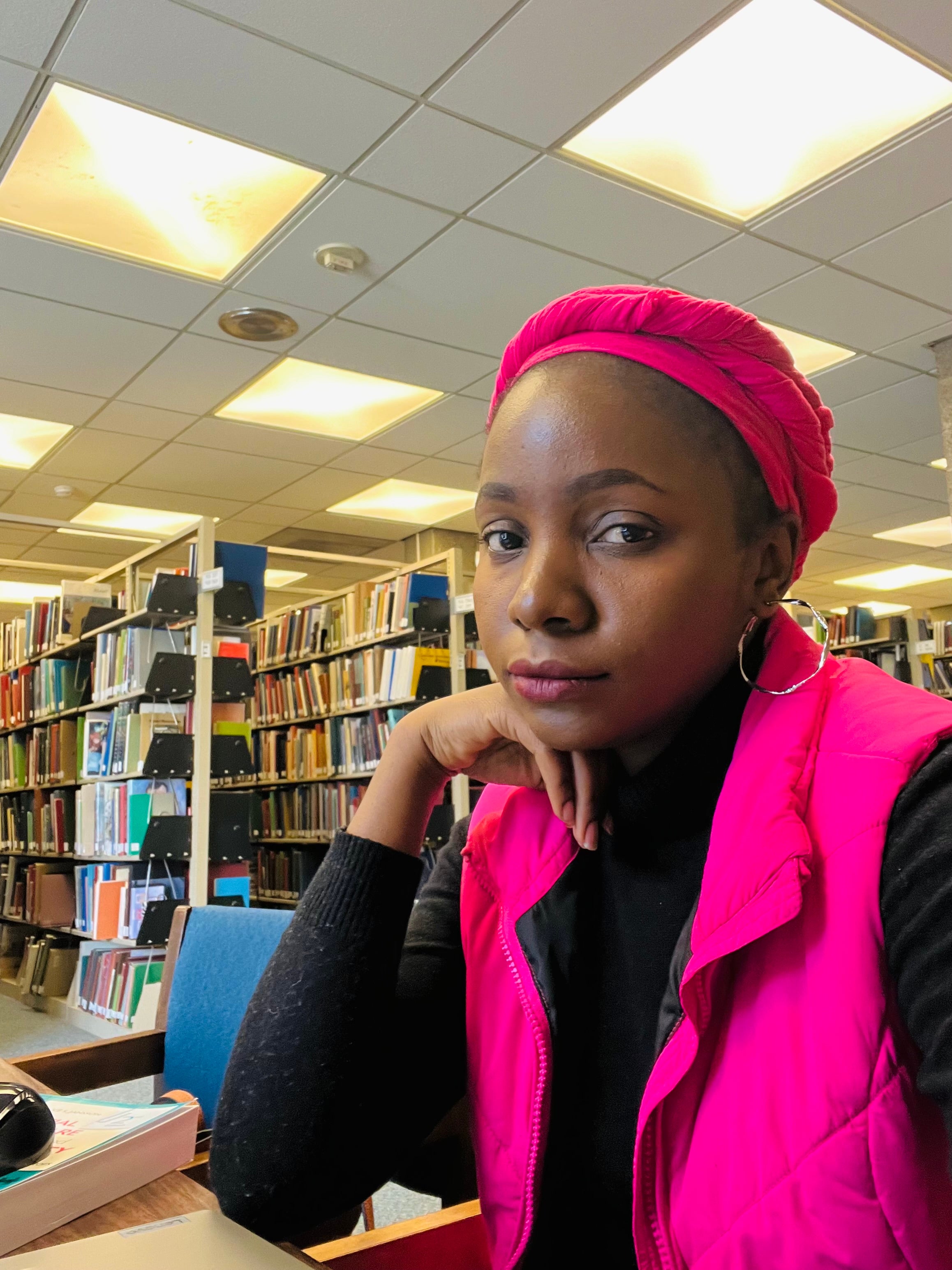 A young Black woman wearing a bright pink headwrap and matching vest sits in a library, resting her chin on her hand. She wears a black long-sleeve top and hoop earrings, with bookshelves and ceiling lights in the background.