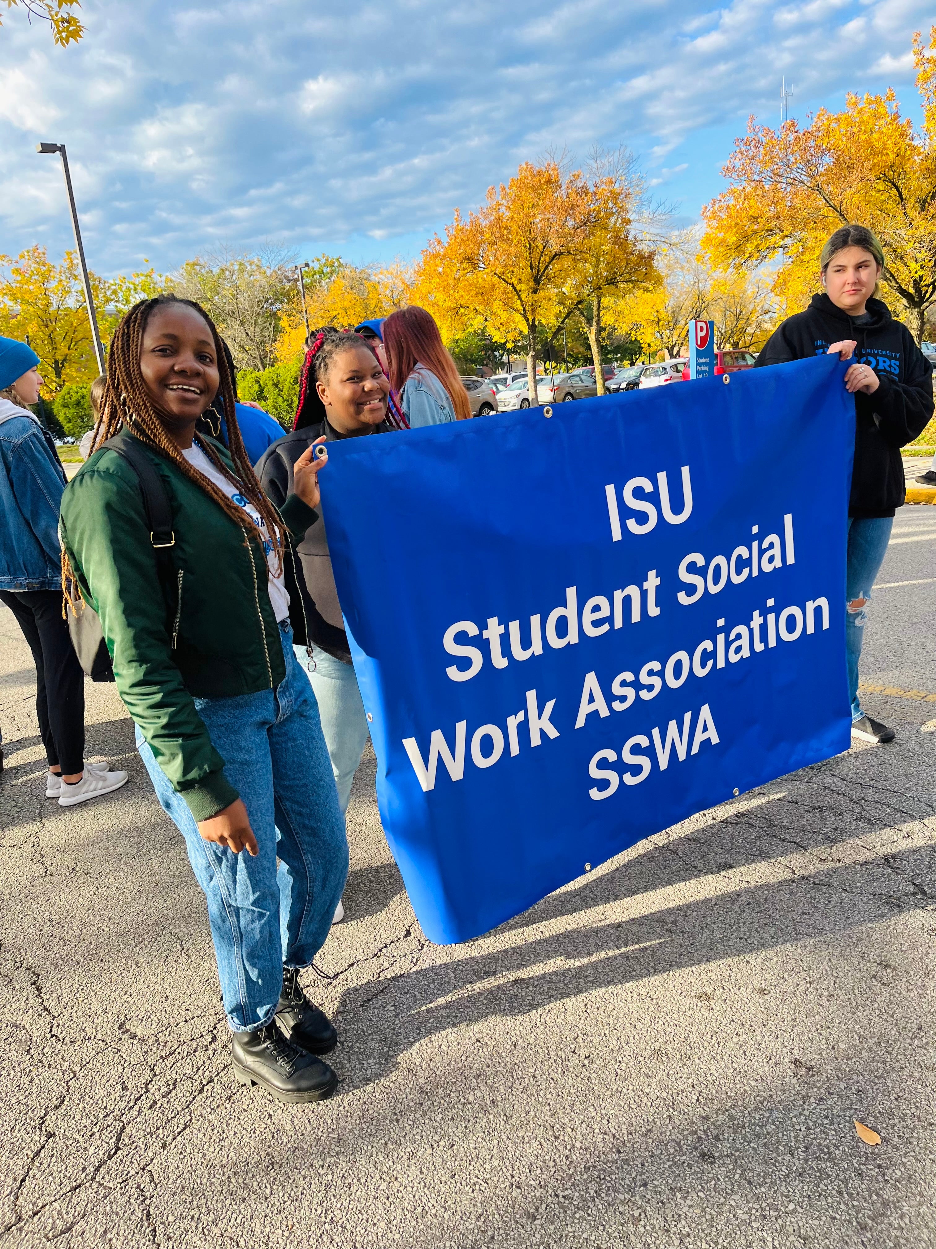 Three young women stand outdoors holding a blue banner that reads “ISU Student Social Work Association SSWA.” The woman on the left, who is Black, smiles while wearing a green jacket, jeans, and black boots.