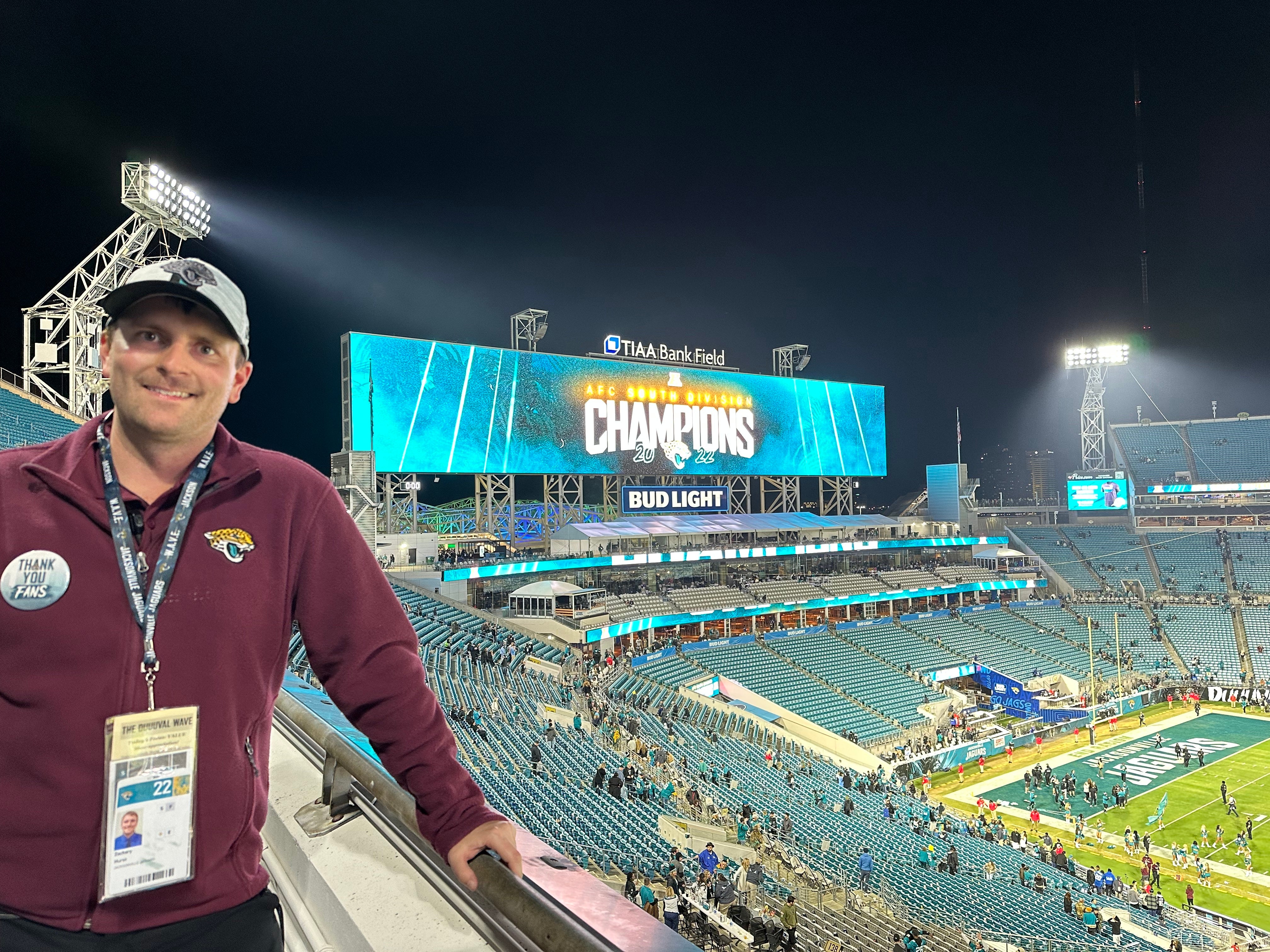 A White man wearing a maroon Jacksonville Jaguars jacket and gray cap stands in a football stadium at night. Behind him, a large screen displays “AFC South Division Champions” above the field at TIAA Bank Field. The stands are partially filled with fans as stadium lights illuminate the scene