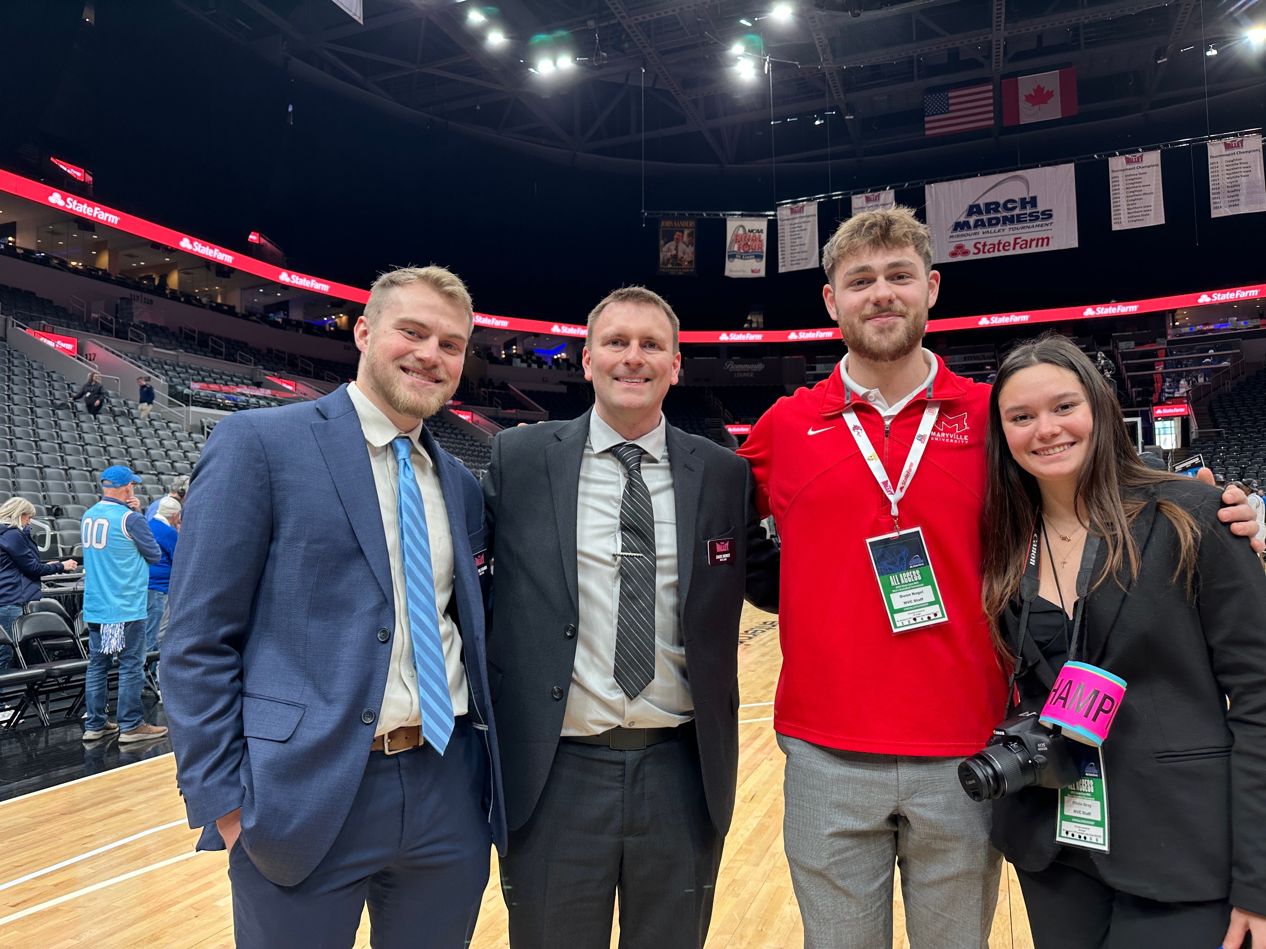 Three White men and a White woman stand courtside at a basketball arena, smiling for a photo. Two men wear suits, one wears a red shirt, and the woman holds a camera with an event badge. Behind them, banners for “Arch Madness” and “State Farm” hang above partially filled stands.
