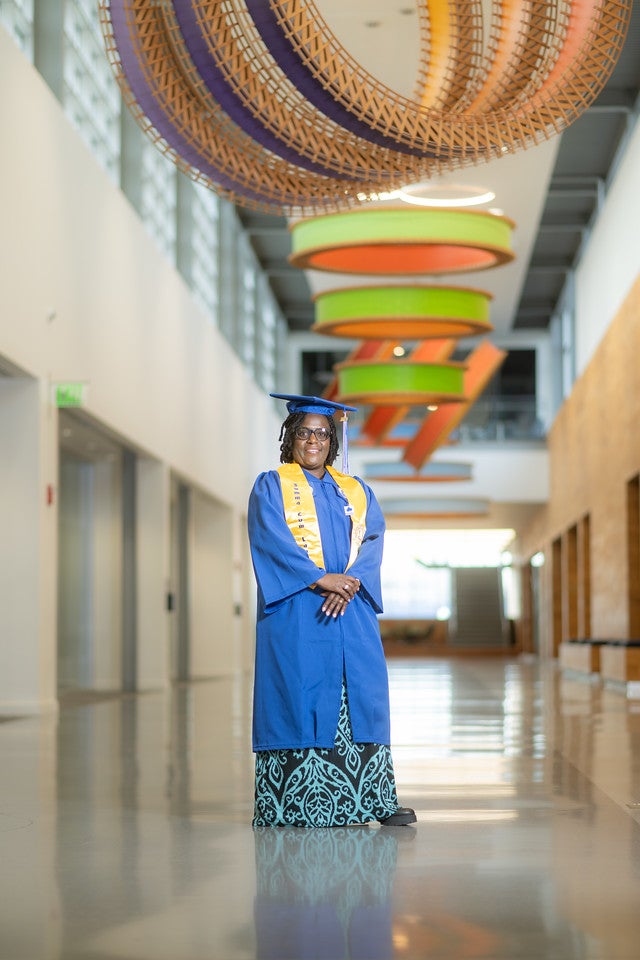 A Black woman wearing a blue graduation gown, yellow stole, and cap stands in a bright hallway with her hands clasped. She smiles softly, and colorful circular ceiling decorations hang above her.