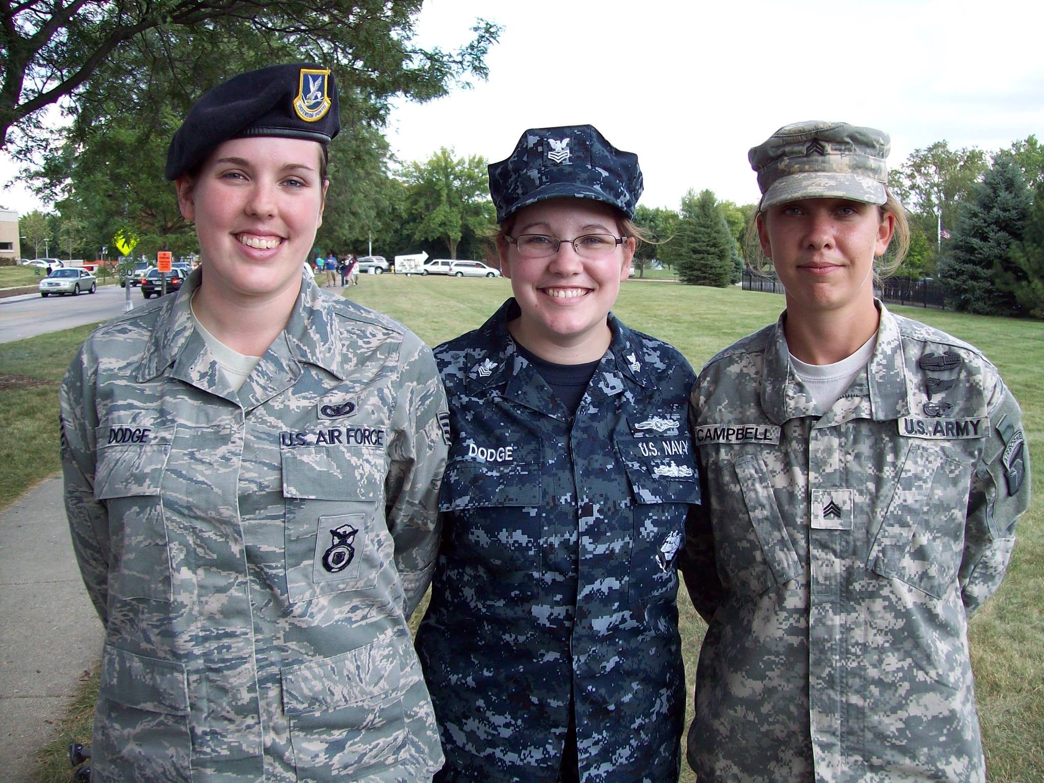 Three individuals standing outdoors in military uniforms representing different branches: U.S. Air Force on the left, U.S. Navy in the center, and U.S. Army on the right. They are positioned side by side on a grassy area with trees and a sidewalk in the background.