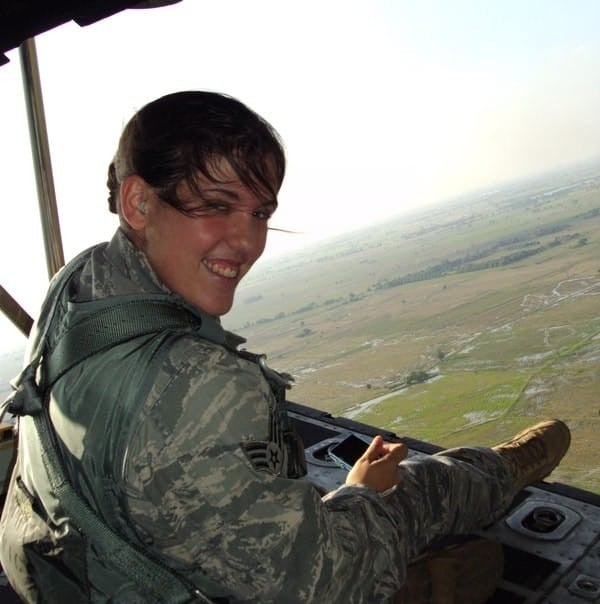 Woman in a military flight uniform seated near an open aircraft door, overlooking a vast landscape of fields and waterways below.