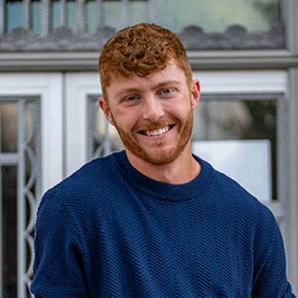 A smiling young man with short red hair and beard, and wearing a blue long-sleeved shirt, stands in front of ornate metal doors. 