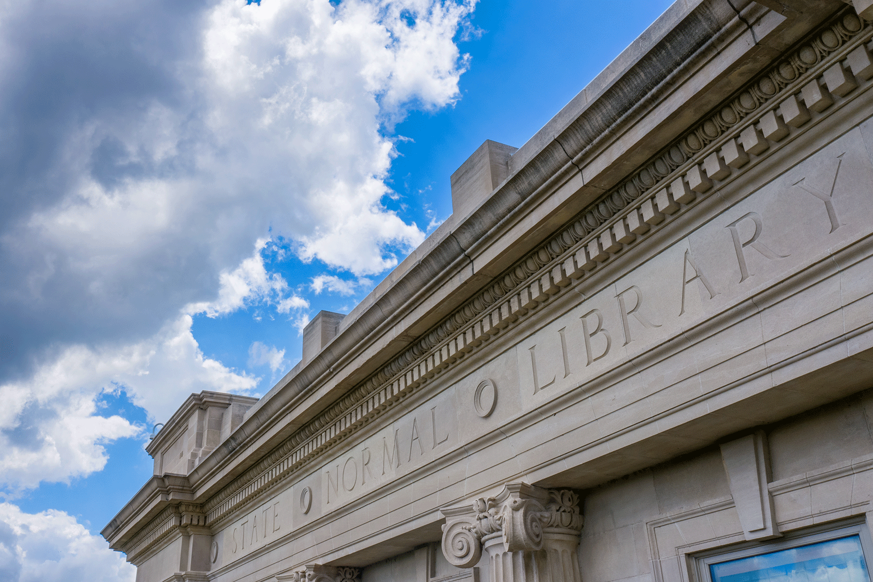 Upper exterior of a building made of light-colored stone, featuring ornate columns and carvings. “State Normal Library” is carved into the building. A partly cloudy blue sky is visible in the background. 