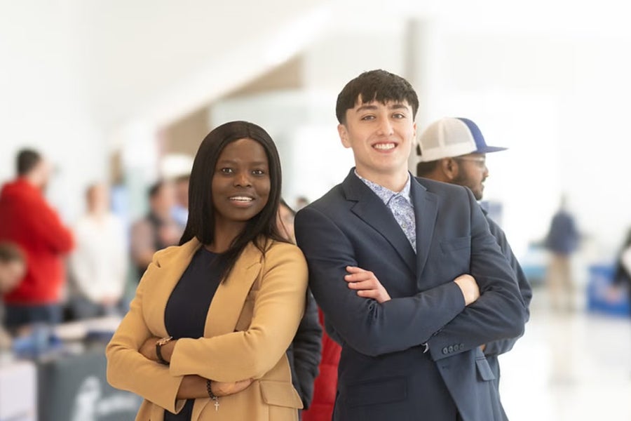 Two individuals standing side by side with arms crossed in a bright indoor setting, dressed in professional attire—a tan blazer over a dark top, and a dark suit with a light shirt. Blurred figures, tables, and banners are in the background, suggesting a career fair or networking event.
