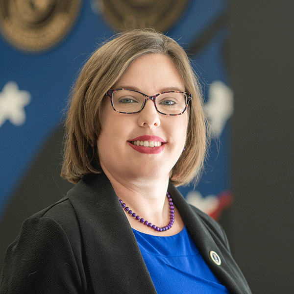 A white woman with chin-length light brown hair and patterned eyeglasses, wearing a black blazer with a small pin on her lapel, a bright blue top, and a purple beaded necklace, smiles while standing indoors. The background features a blurred blue and black mural with white star shapes.