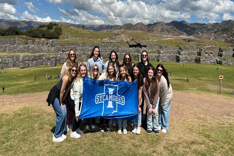 Group of people posing outdoors in front of ancient stone terraces. They hold a blue banner with a white “I” and the word “Sycamores” representing Indiana State University. Green fields and distant mountains are visible behind them.