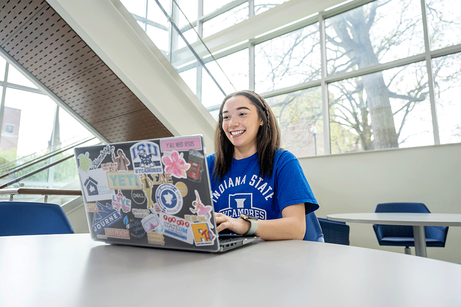 Female student with dark brown hair sitting at a table in a bright indoor space with large windows, using a laptop covered in colorful stickers. She is wearing a blue T-shirt that reads “Indiana State Sycamores” with the university logo. 