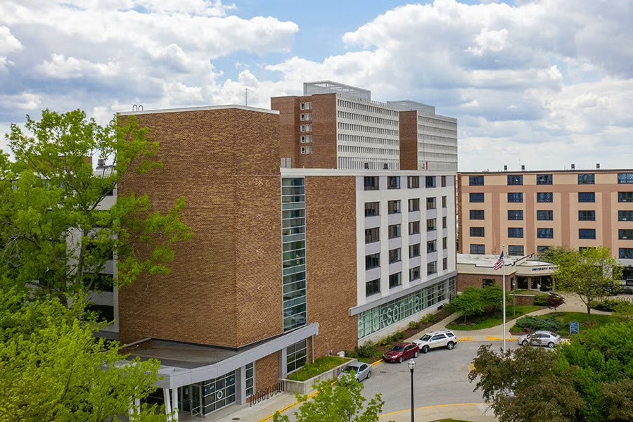 A large multi-story building complex with brown brick and glass panels in the foreground and taller structures visible behind. Cars are parked along a curved driveway near the entrance. Green trees and landscaped areas surround the buildings under a partly cloudy sky. 