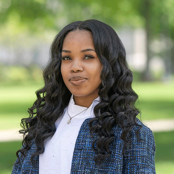 A young Black woman with long, wavy dark hair stands outdoors, wearing a white blouse and a blue textured jacket. She has a small nose ring and a delicate necklace. The background shows a softly blurred green scenery.