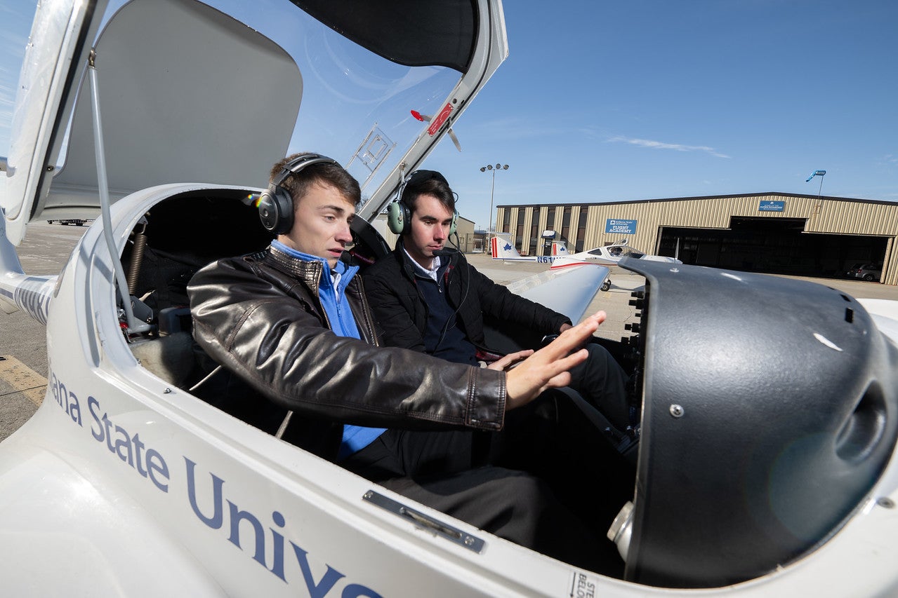 Two white males seated inside a small aircraft with the canopy open, both wearing headsets. The person in the front left seat extends a hand toward the aircraft controls, while a hangar and additional planes are visible in the background.