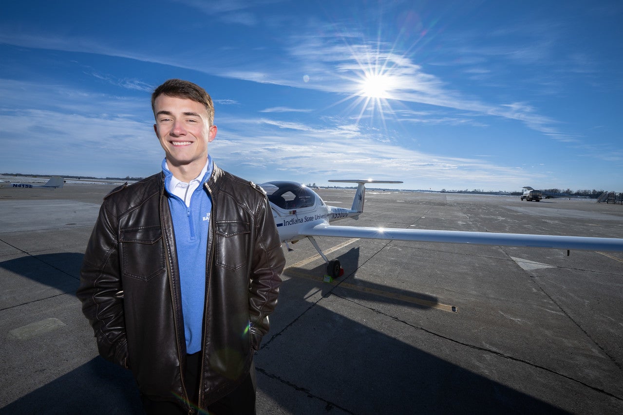 A white male with short brown hair wearing a brown leather jacket and a blue quarter‑zip sweater stands on an airport tarmac with a small white aircraft behind them. The sun shines brightly in a clear blue sky.