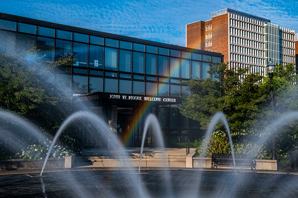 Arched streams of water from a fountain frame the John W. Moore Welcome Center, with a clear rainbow appearing in front of the building’s glass façade. Trees surround the entrance, and a tall brick campus building stands in the background under a blue sky.