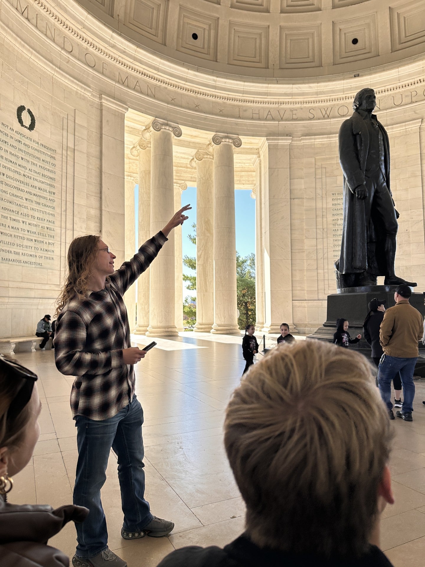 Inside the Jefferson Memorial, a person in a plaid shirt stands with one arm raised, gesturing upward. Several people observe from nearby, and the large bronze statue of Thomas Jefferson towers in the background. Sunlight streams through the tall marble columns surrounding the memorial’s interior.