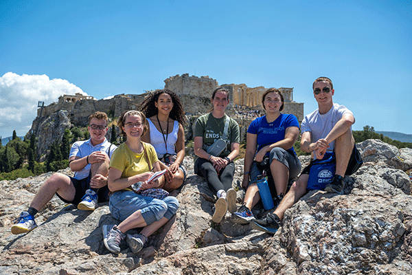 A group of six people sit on rocky terrain with the ancient Acropolis ruins visible in the background under a clear blue sky. They are casually dressed and appear to be resting or posing together, with trees and distant hills adding to the scenic outdoor setting.