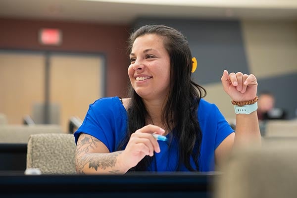 A smiling woman with long black hair sits at a table in a classroom or meeting space, wearing a blue top and holding a pen, with a laptop or desk surface in the foreground. The student appears happy and engaged in discussion, with an exit sign and other people visible in the softly lit background.