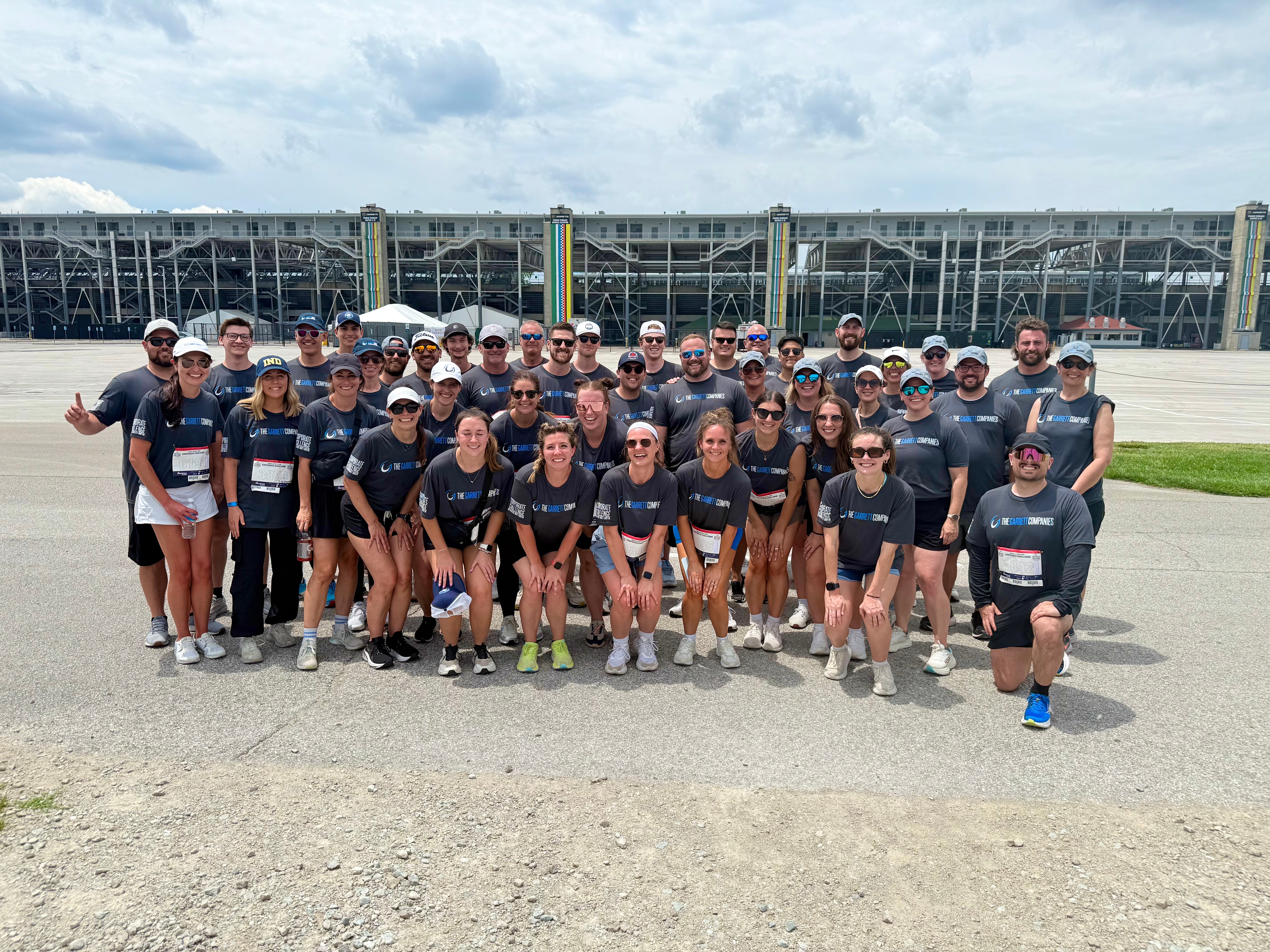 Group of people wearing matching dark T-shirts pose together outdoors on a paved area, with a large stadium structure visible in the background under a partly cloudy sky.