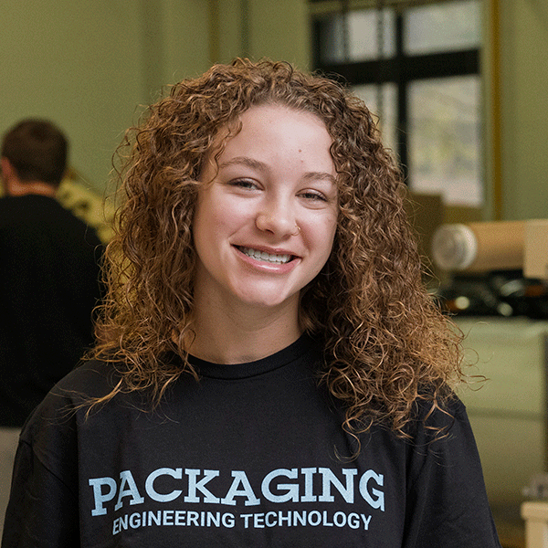 “Smiling young woman with curly hair wearing a black ‘Packaging Engineering Technology’ sweatshirt stands indoors with a blurred background.”