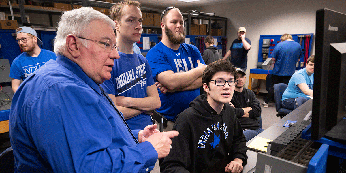 A group of people in an engineering lab gather around a computer workstation. An instructor in a blue shirt gestures toward the screen while students in Indiana State University apparel observe and work at nearby stations.