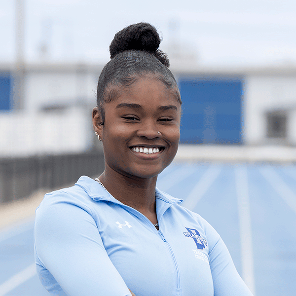 Smiling woman wearing a light blue athletic jacket stands on an outdoor track with stadium seating blurred in the background.