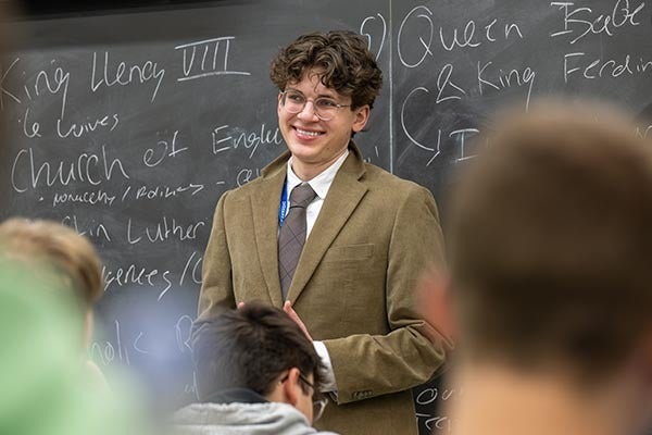 “Young man wearing glasses and a brown blazer smiles while standing in front of a chalkboard with handwritten notes in a classroom.”