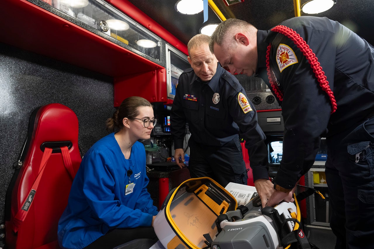 Inside an ambulance, two emergency responders stand beside a seated healthcare worker in blue scrubs, reviewing medical equipment and documentation in a brightly lit, red‑and‑black interior.