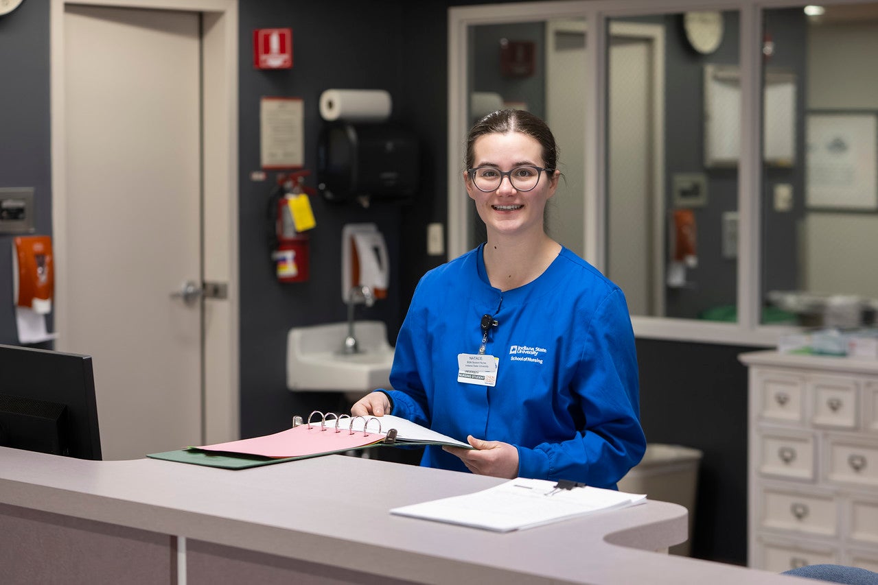 A white female student with brown hair pulled back into a ponytail wears blue nursing scrubs. She stands behind a reception desk in a healthcare setting holding an open binder with documents. Sinks, cabinets, and medical supplies are visible in the background.