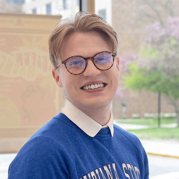 Smiling man wearing glasses and a blue Indiana State sweatshirt stands indoors near a window with a blurred campus view outside.