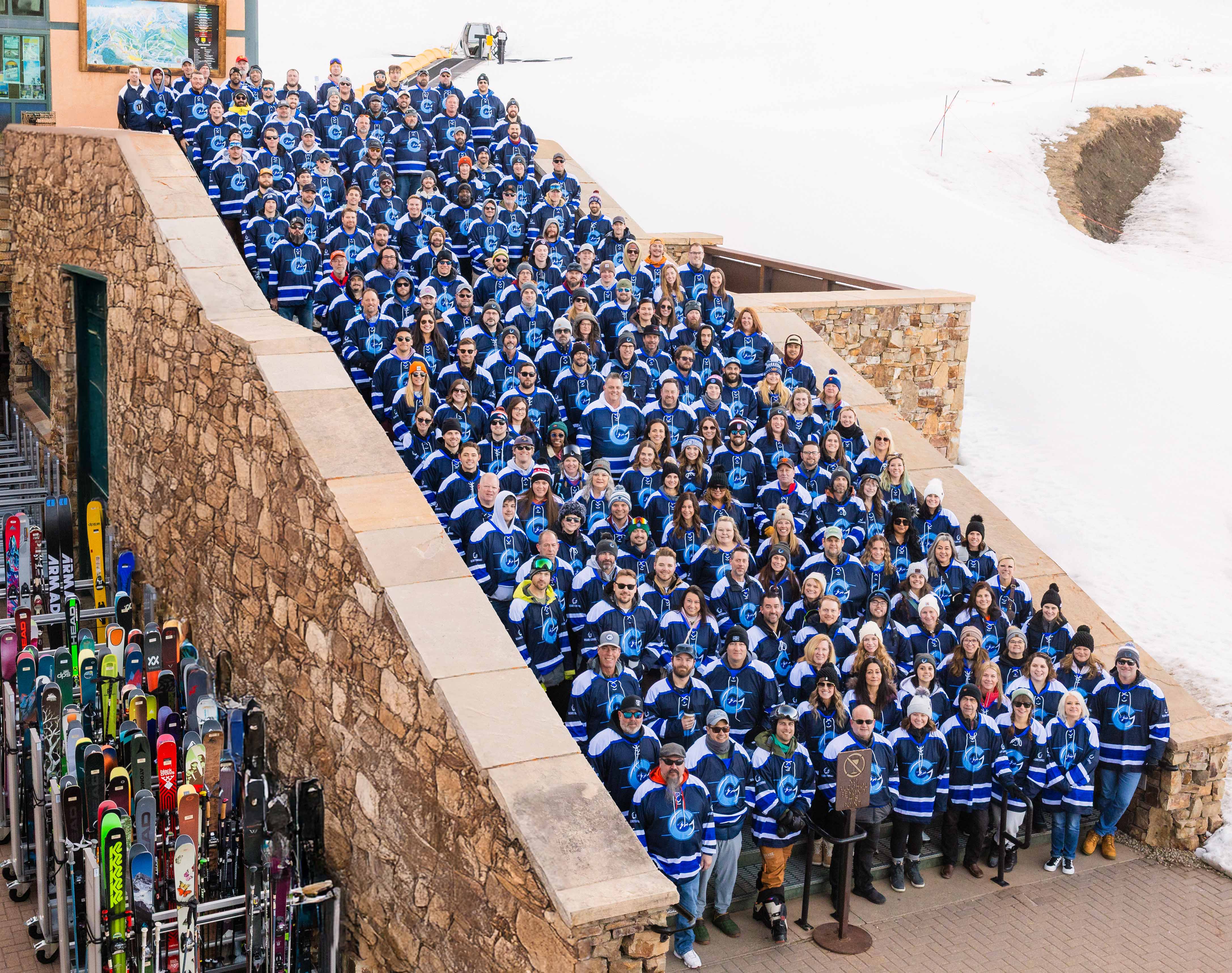 Large group of people wearing matching blue skiing outfits gathered on outdoor stone steps at a snowy mountain resort, with ski racks and equipment visible along the side.