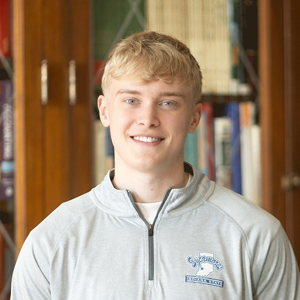 “Smiling young man wearing a light gray Indiana State sweatshirt stands in front of bookshelves indoors.”