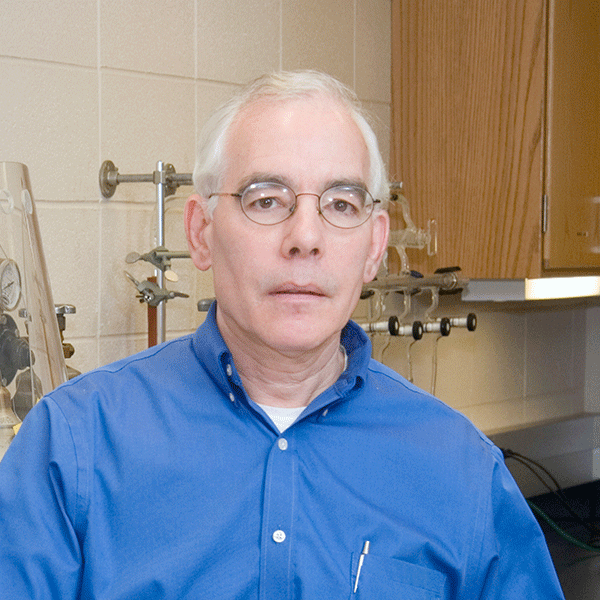 Man with white hair and glasses wearing a blue button-down shirt stands in a laboratory with equipment in the background.
