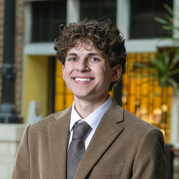 Smiling young man with curly brown hair, wearing a tan suit jacket, white dress shirt, and patterned tie, stands indoors in a modern setting with large windows, warm ambient lighting, and greenery in the background.