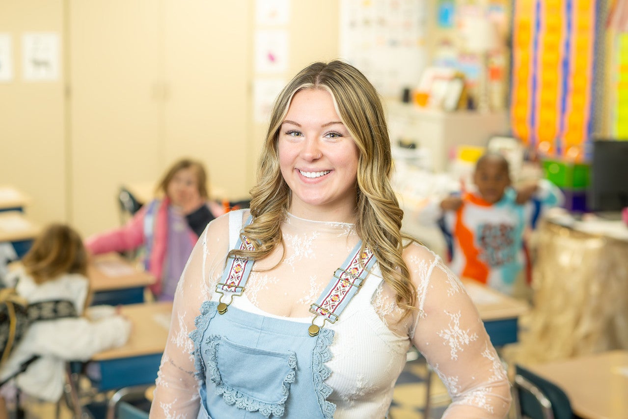 A white female student with wavy blond hair stands at the front of an elementary classroom, with children seated at desks and colorful educational posters visible in the background. The student wears a white lace long-sleeved shirt with light blue denim overalls. 