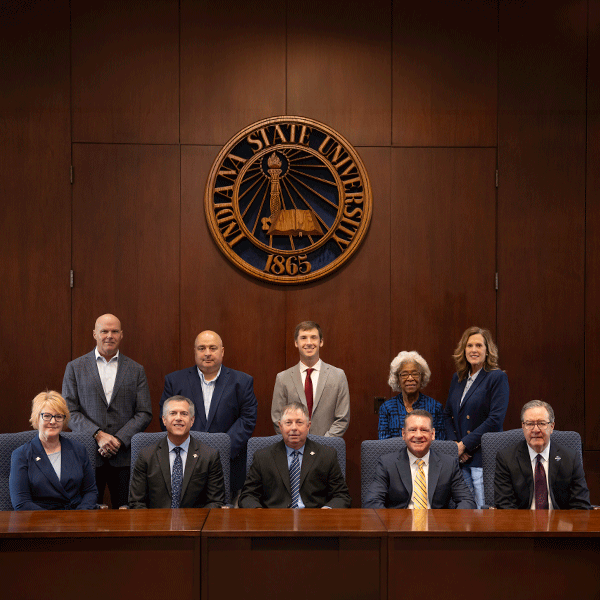 A group of formally dressed university leaders seated and standing in a wood-paneled boardroom beneath the Indiana State University seal, posed for an official group portrait.