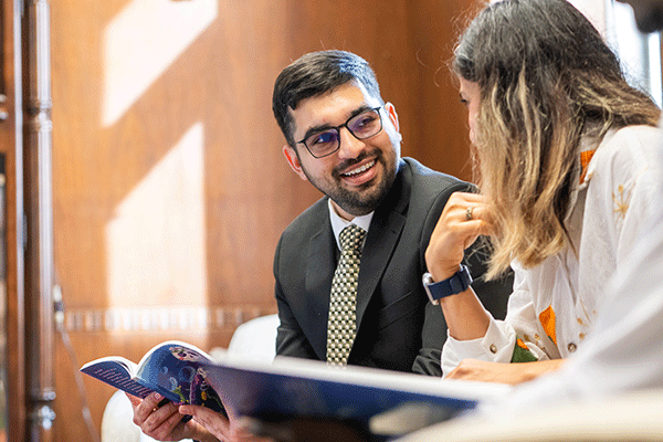 Two people seated indoors review a booklet together, with one person in business attire and the other gesturing while discussing the material in a wood-paneling setting