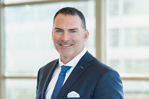 A professionally dressed man with short, dark hair and wearing a navy suit, white shirt, and patterned blue tie stands indoors in front of large windows, with soft natural light illuminating the background.